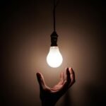 A hand reaching towards a glowing hanging light bulb symbolizing creativity and illumination.